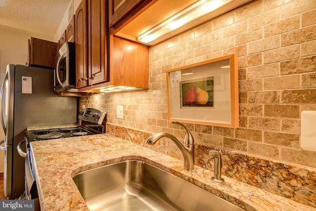 kitchen with stainless steel appliances, brown cabinetry, a sink, a textured ceiling, and light stone countertops