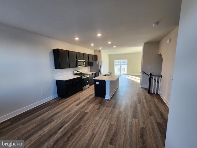 kitchen with dark wood finished floors, an island with sink, stainless steel appliances, dark cabinetry, and light countertops