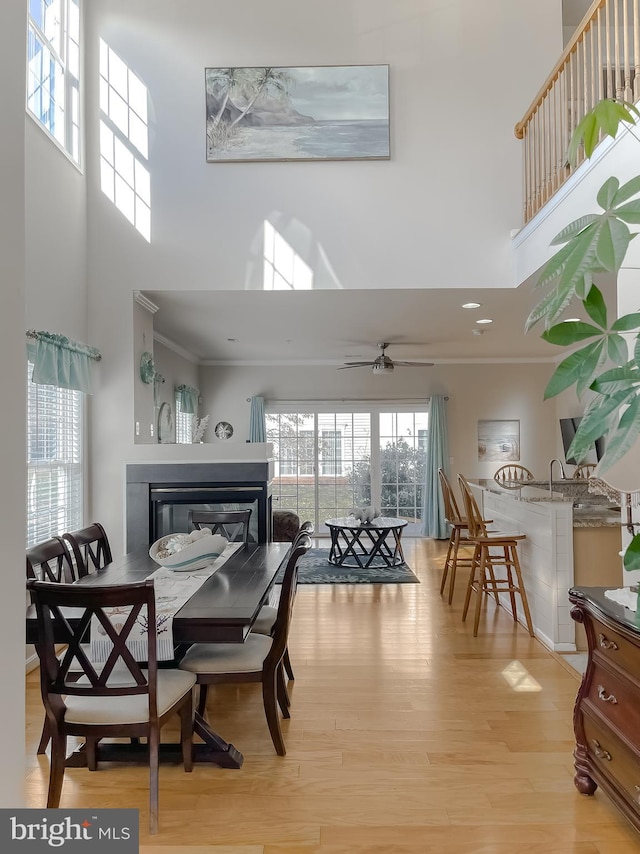 dining room with light wood finished floors, a towering ceiling, ornamental molding, a glass covered fireplace, and a ceiling fan