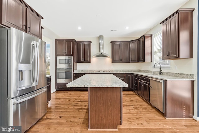 kitchen featuring stainless steel appliances, a sink, light wood-style floors, a center island, and wall chimney exhaust hood