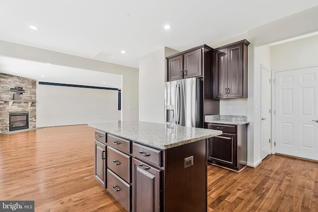 kitchen with a kitchen island, light stone counters, dark brown cabinets, a stone fireplace, and stainless steel refrigerator with ice dispenser