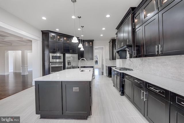 kitchen featuring a sink, hanging light fixtures, appliances with stainless steel finishes, an island with sink, and glass insert cabinets