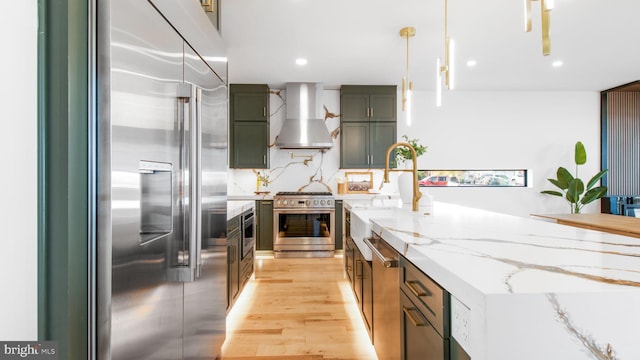 kitchen featuring high end appliances, a sink, wall chimney exhaust hood, light stone countertops, and light wood-style flooring