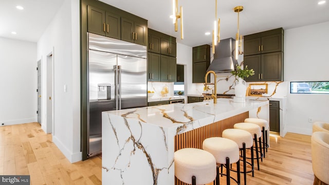 kitchen with recessed lighting, light wood-type flooring, light stone countertops, wall chimney exhaust hood, and stainless steel built in refrigerator