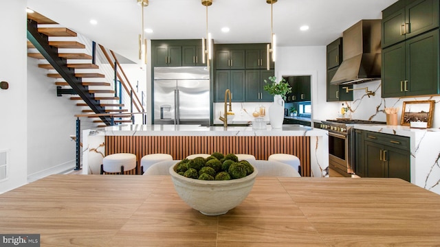 kitchen featuring high end appliances, green cabinetry, light countertops, wall chimney range hood, and a sink