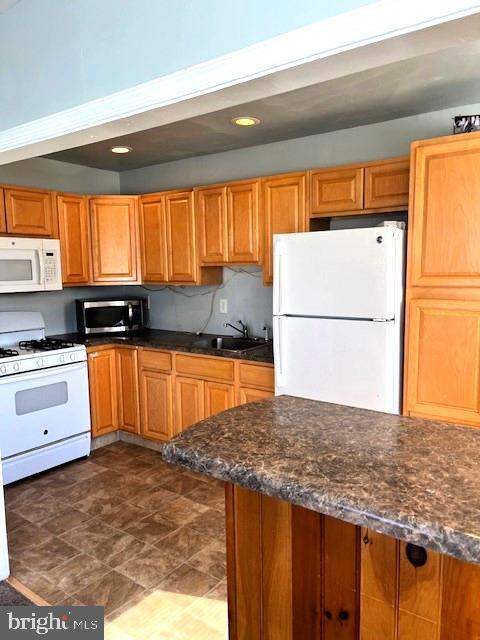 kitchen with recessed lighting, brown cabinetry, a sink, dark stone countertops, and white appliances