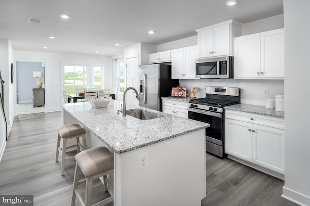 kitchen with white cabinets, appliances with stainless steel finishes, light stone counters, a kitchen island with sink, and a sink