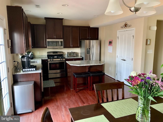 kitchen with dark brown cabinetry, stainless steel appliances, a center island, light stone countertops, and dark wood-style floors