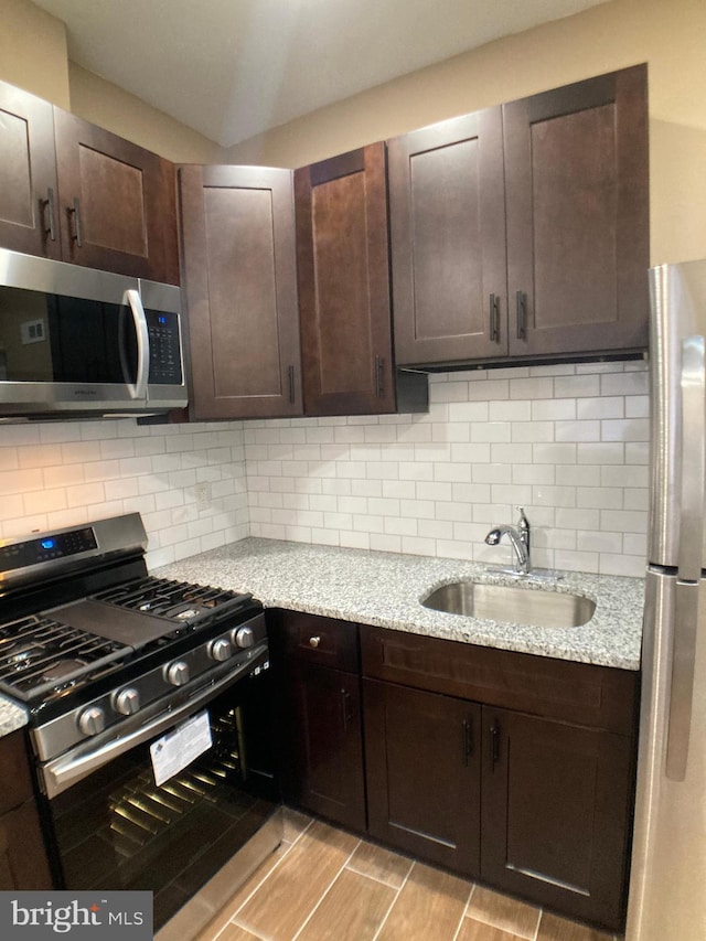 kitchen with tasteful backsplash, appliances with stainless steel finishes, a sink, dark brown cabinets, and light stone countertops