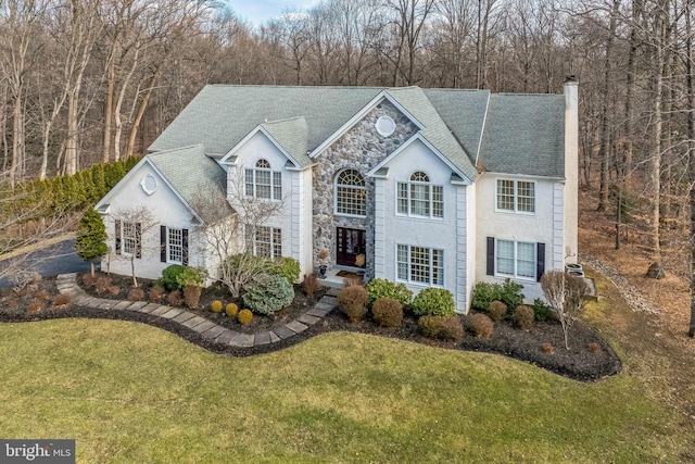 view of front of home featuring stone siding, a chimney, a wooded view, and a front yard