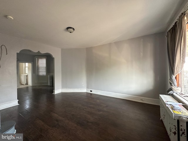 unfurnished room featuring dark wood-type flooring, arched walkways, radiator, and baseboards