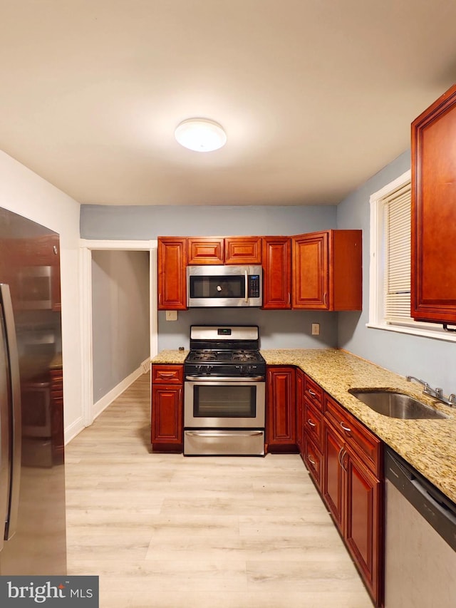 kitchen with reddish brown cabinets, appliances with stainless steel finishes, light wood-style floors, a sink, and light stone countertops