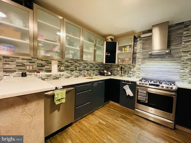 kitchen with stainless steel appliances, light countertops, glass insert cabinets, light wood-type flooring, and wall chimney exhaust hood