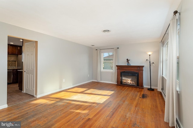 unfurnished living room with light wood-style flooring, visible vents, baseboards, baseboard heating, and a glass covered fireplace