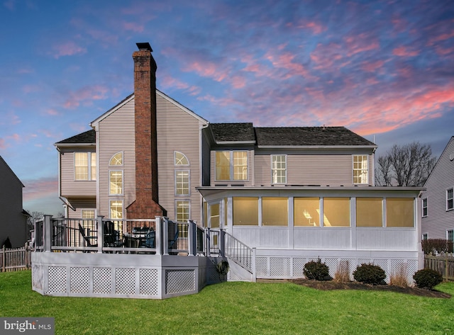 back of house at dusk featuring a yard, a chimney, a wooden deck, and a sunroom