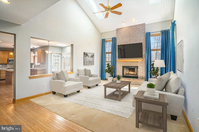 living room with baseboards, ceiling fan, light wood-type flooring, a fireplace, and high vaulted ceiling