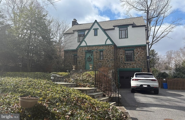 tudor home with driveway, a garage, a chimney, fence, and stucco siding