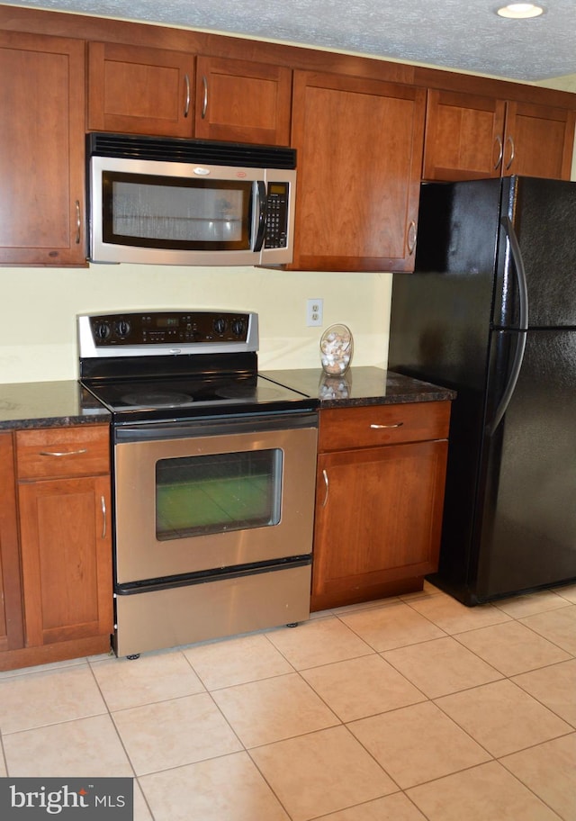 kitchen with brown cabinetry, light tile patterned floors, stainless steel appliances, and dark stone countertops