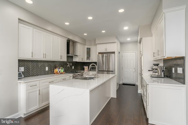 kitchen featuring an island with sink, appliances with stainless steel finishes, wall chimney range hood, white cabinetry, and a sink