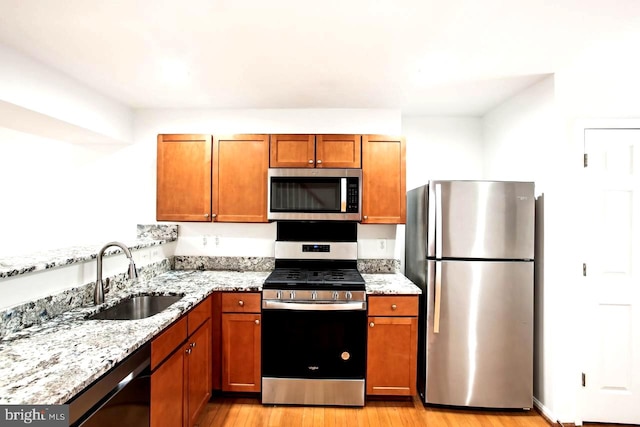 kitchen with brown cabinetry, light wood-style flooring, appliances with stainless steel finishes, light stone counters, and a sink