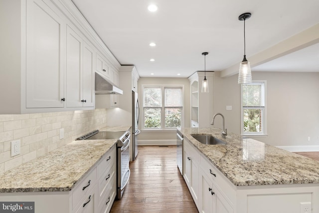 kitchen featuring an island with sink, hanging light fixtures, stainless steel appliances, under cabinet range hood, and a sink