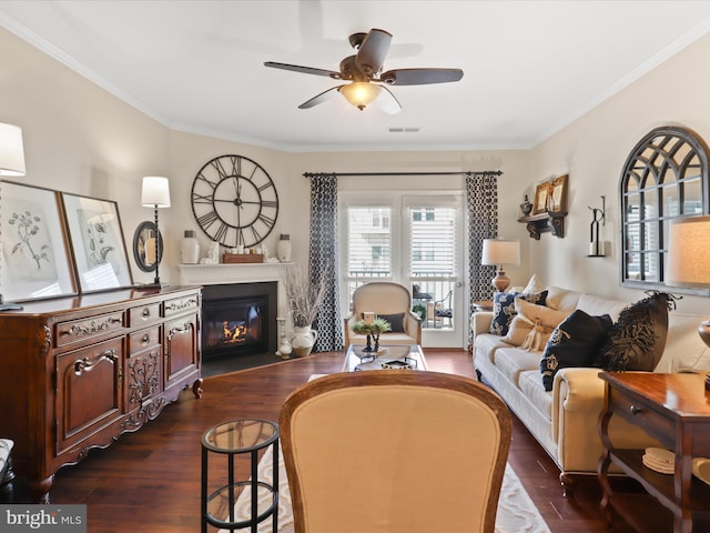 living area featuring crown molding, dark wood finished floors, visible vents, a glass covered fireplace, and ceiling fan