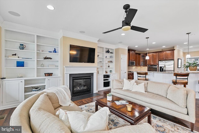 living area with dark wood finished floors, a ceiling fan, a fireplace with flush hearth, crown molding, and recessed lighting