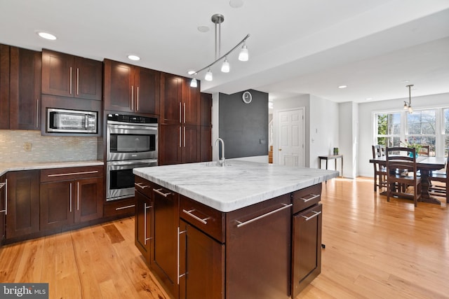 kitchen with light wood finished floors, appliances with stainless steel finishes, an island with sink, and pendant lighting