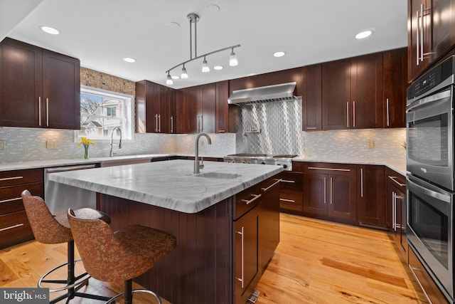 kitchen featuring a kitchen island with sink, stainless steel appliances, light wood-type flooring, wall chimney range hood, and a kitchen bar