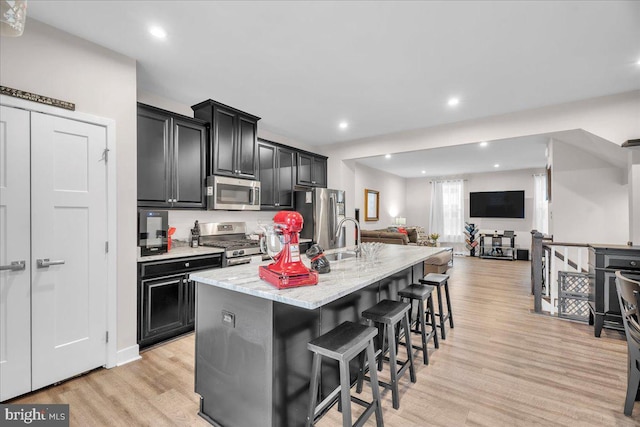 kitchen featuring stainless steel appliances, light wood-style flooring, open floor plan, a kitchen island with sink, and dark cabinets