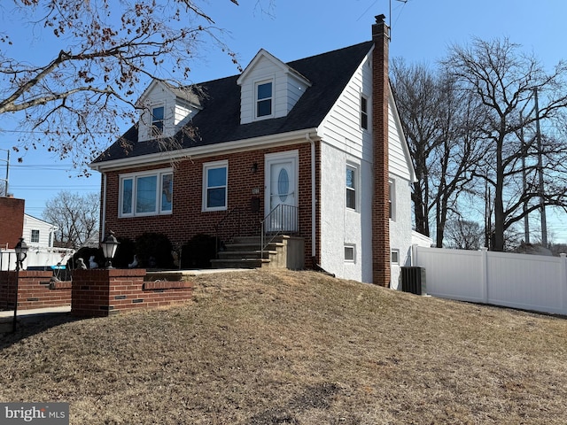 new england style home featuring brick siding, a chimney, central AC unit, fence, and a front lawn