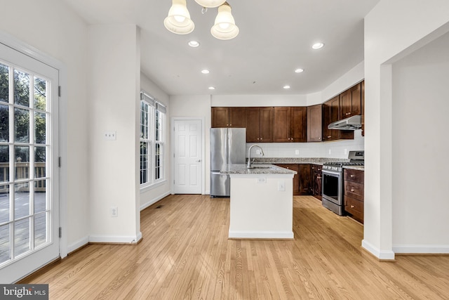 kitchen featuring under cabinet range hood, light wood-style floors, appliances with stainless steel finishes, light stone countertops, and a center island with sink