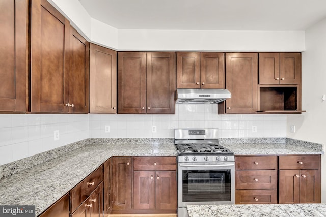 kitchen with under cabinet range hood, light stone countertops, stainless steel gas stove, tasteful backsplash, and brown cabinetry