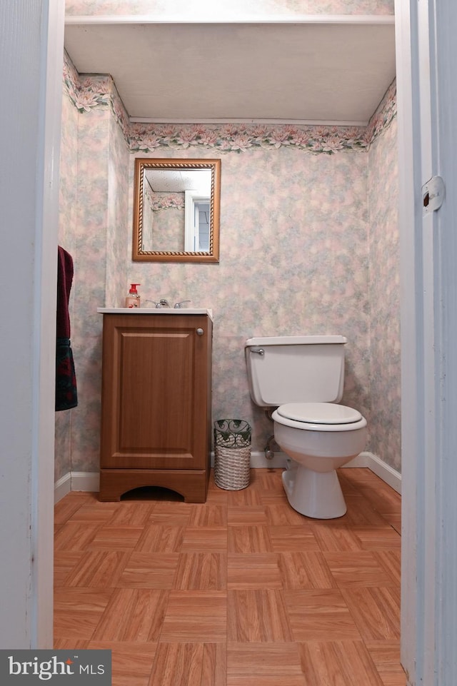 half bath with baseboards, toilet, vanity, and wallpapered walls