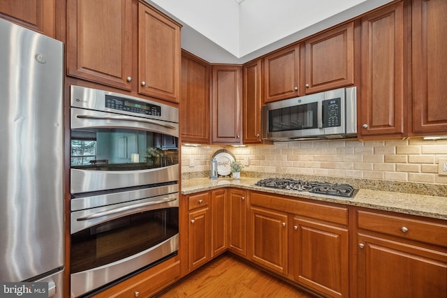 kitchen with brown cabinetry, light wood-style flooring, appliances with stainless steel finishes, light stone countertops, and backsplash