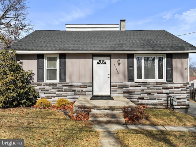 view of front facade with stone siding, a shingled roof, a chimney, and a front yard