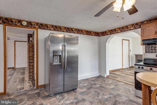 kitchen featuring baseboards, arched walkways, appliances with stainless steel finishes, a textured ceiling, and under cabinet range hood