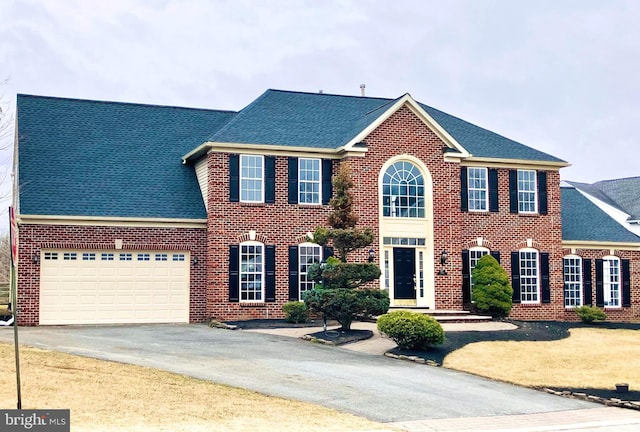 colonial house with driveway, roof with shingles, a garage, and brick siding