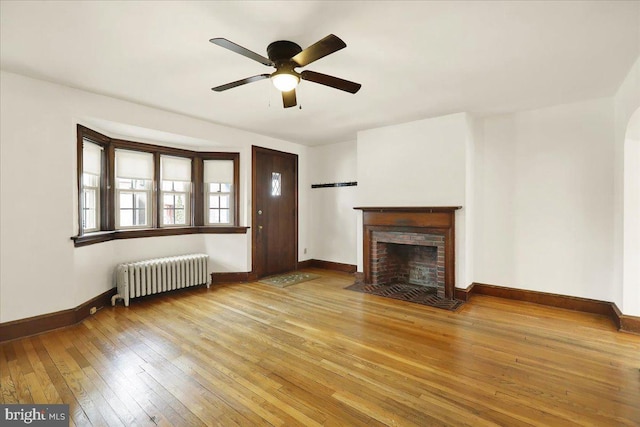 unfurnished living room with baseboards, a ceiling fan, radiator heating unit, light wood-style flooring, and a brick fireplace