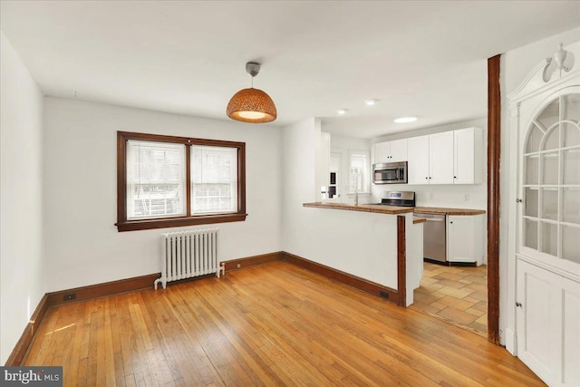 kitchen featuring stainless steel appliances, a peninsula, white cabinets, radiator heating unit, and pendant lighting