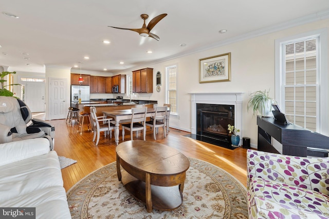 living area with light wood-style floors, a fireplace with flush hearth, ornamental molding, and a ceiling fan