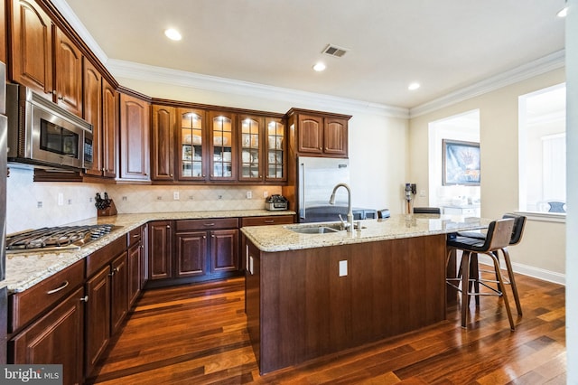 kitchen with an island with sink, appliances with stainless steel finishes, glass insert cabinets, a sink, and backsplash