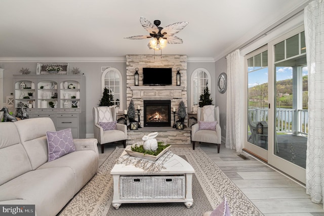 living area with ornamental molding, light wood-style flooring, a stone fireplace, and a ceiling fan