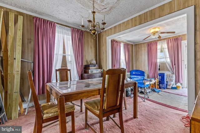 carpeted dining room featuring a textured ceiling, ornamental molding, wood walls, and ceiling fan with notable chandelier