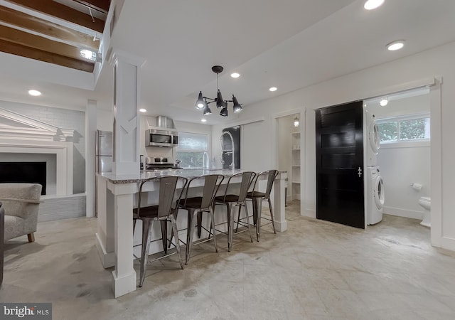 kitchen with light stone counters, stainless steel appliances, white cabinets, stacked washer / drying machine, and a kitchen bar