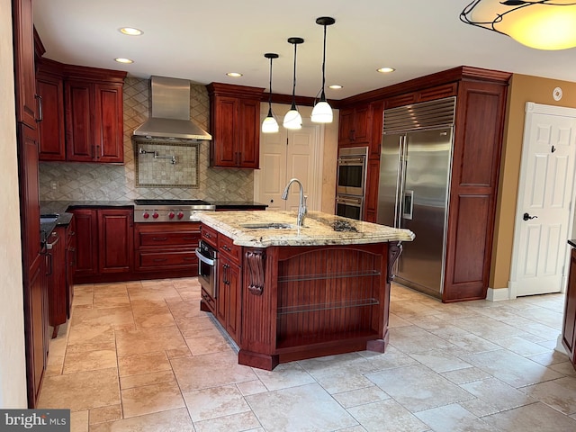 kitchen featuring reddish brown cabinets, wall chimney exhaust hood, light stone countertops, a kitchen island with sink, and stainless steel appliances