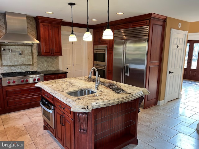 kitchen with an island with sink, appliances with stainless steel finishes, light stone countertops, wall chimney range hood, and a sink