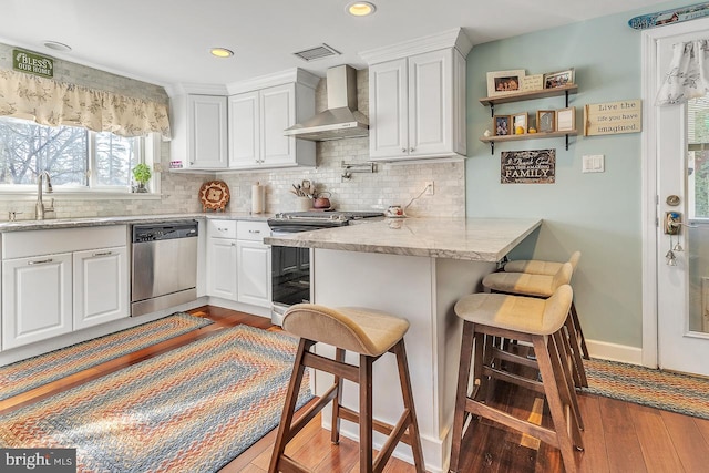 kitchen featuring white cabinets, appliances with stainless steel finishes, a breakfast bar area, wall chimney range hood, and a sink