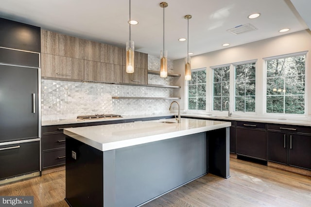 kitchen with paneled refrigerator, a sink, light countertops, open shelves, and an island with sink