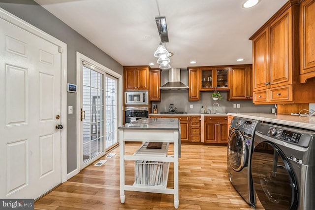 kitchen featuring appliances with stainless steel finishes, brown cabinets, washer and dryer, light wood-type flooring, and wall chimney range hood